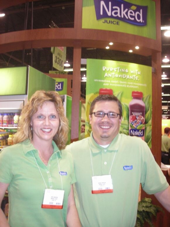 A woman and a man in light green Naked Juice polo shirts and badges smile in front of a Naked Juice banner showing a pomegranate blueberry bottle.