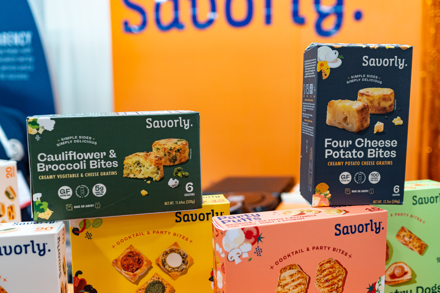 Boxes of Savorly cauliflower and broccoli bites and four cheese potato bites against an orange booth wall