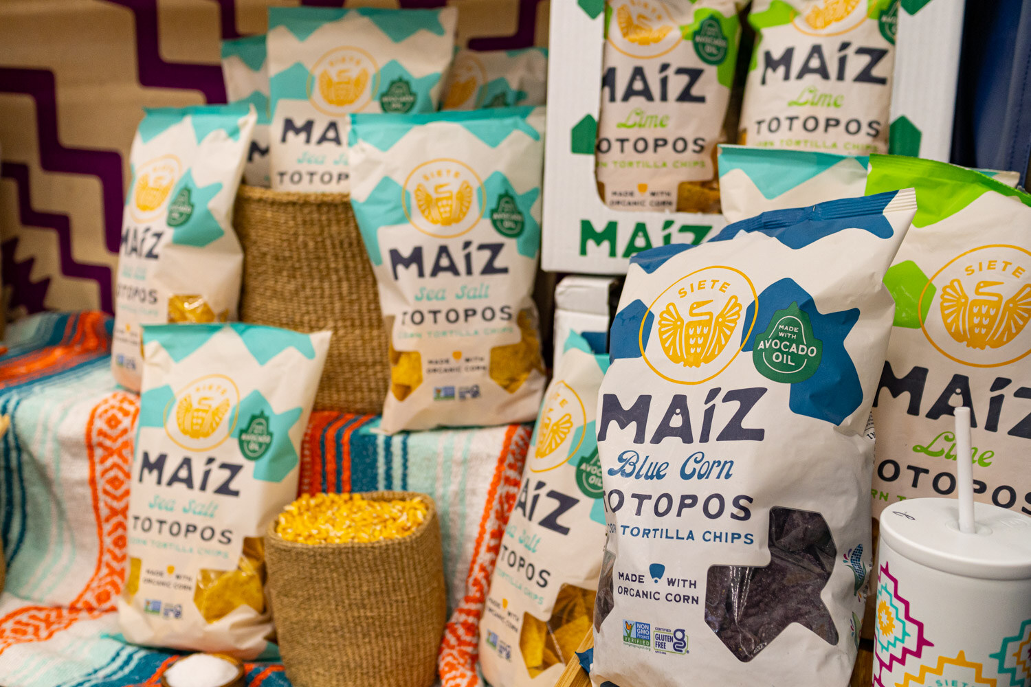 Bags of Maiz Totopos blue corn tortilla chips displayed with baskets of corn