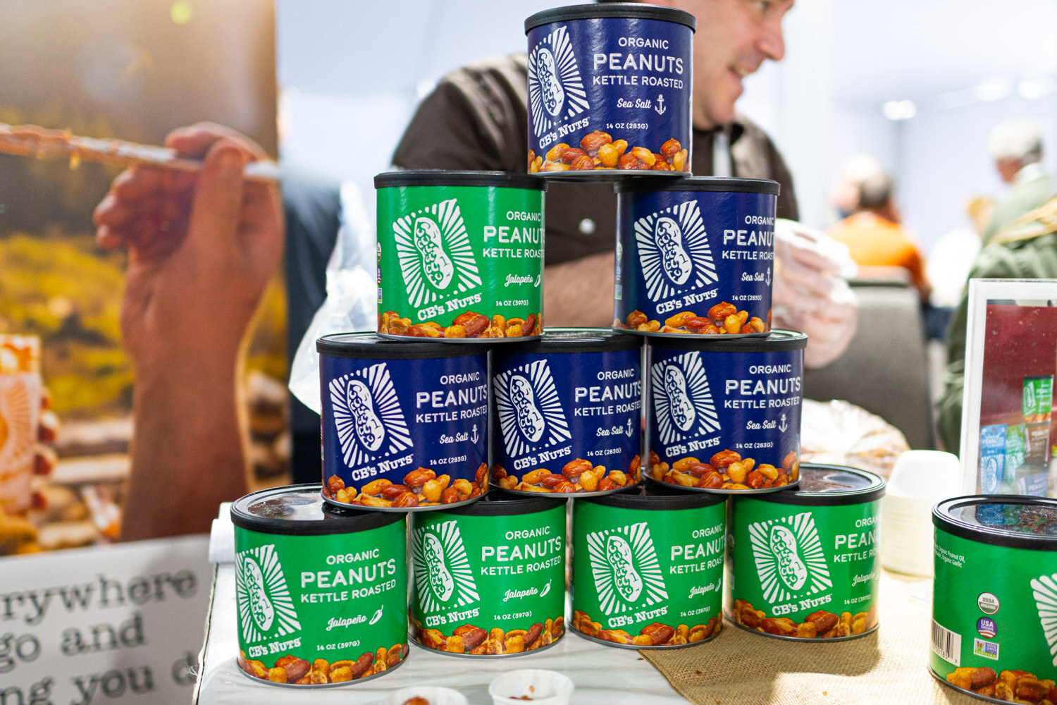 A tall stack of green and blue canned boiled peanuts with a person sampling behind them