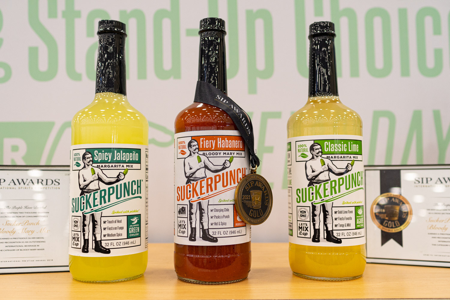 Three Suckerpunch pickle juice bottles in front of a sign reading Stand-Up Choice