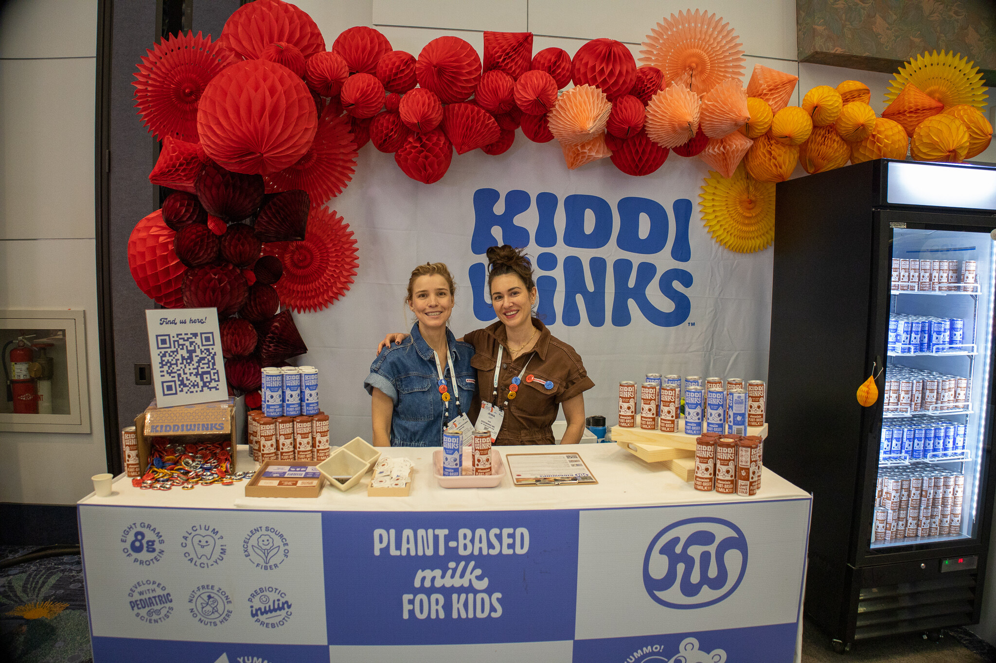 Two booth staff stand behind a Kiddi Drinks counter decorated with red paper flowers