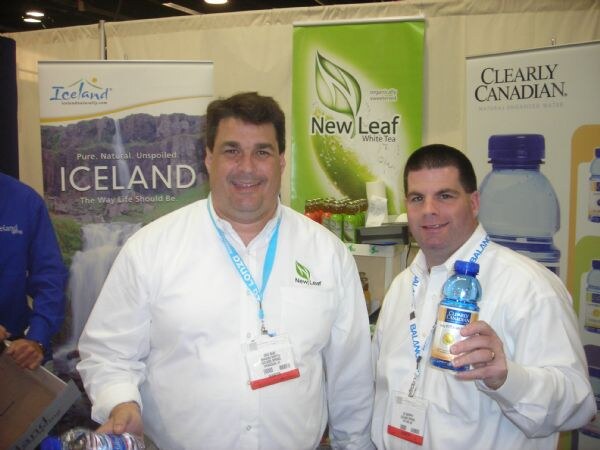 Two men in white shirts stand in front of banners for Iceland, New Leaf white tea and Clearly Canadian. The man on the right holds a Clearly Canadian bottle.