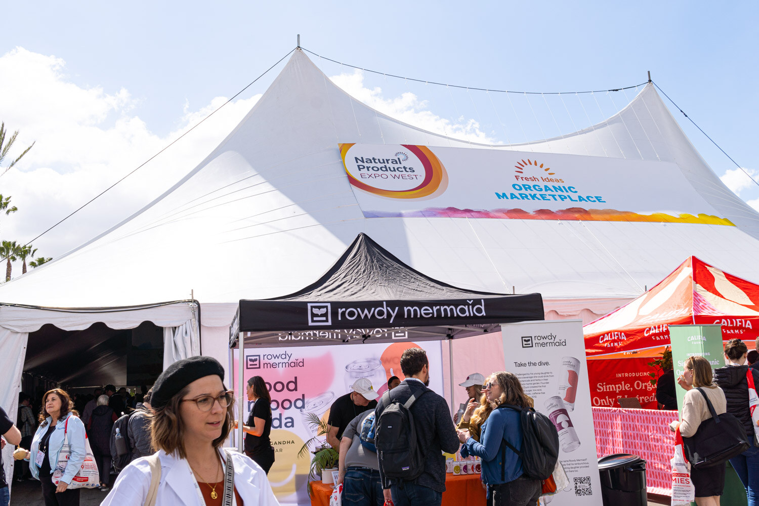 A white tent labeled Natural Marketplaces with attendees and booths in front of it