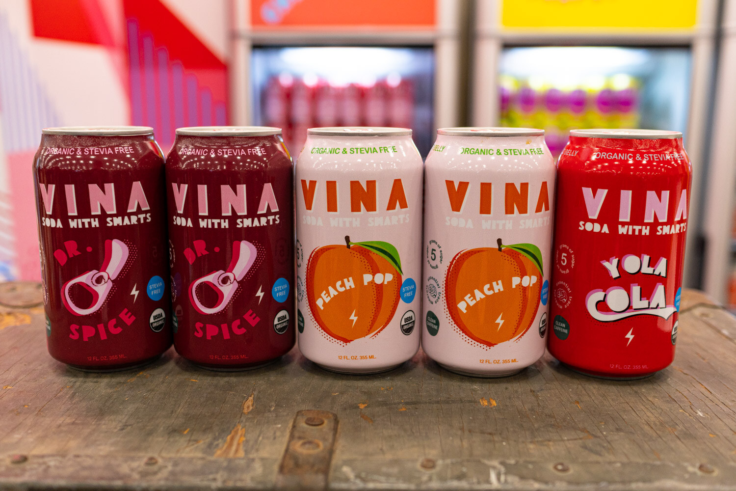 Five Vina soda cans in cherry, peach pop and cola flavors on a wooden board