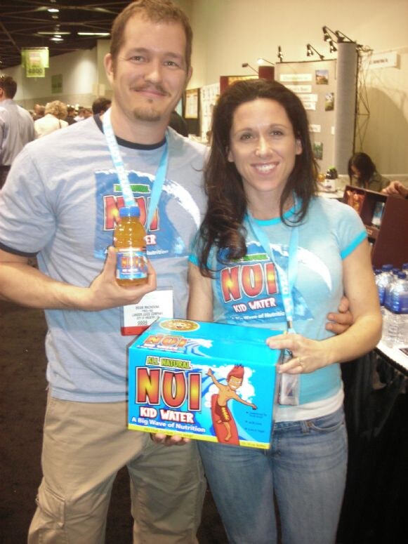 A man and a woman in matching blue Nui Kid Water T-shirts stand together; he holds a small bottle of juice and she holds a box of Nui Kid Water.