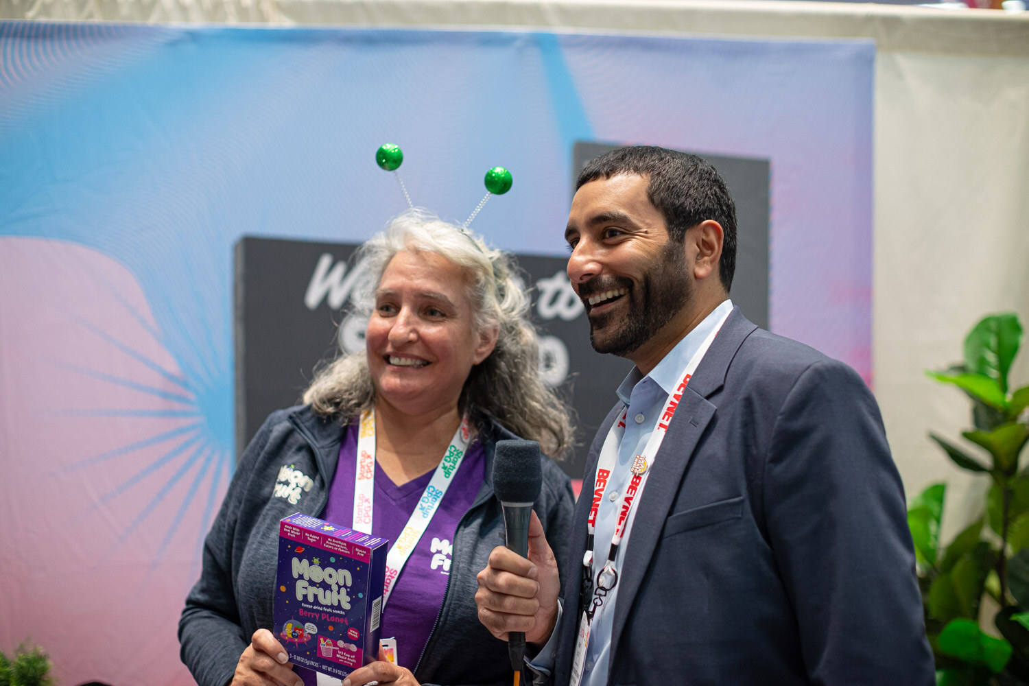 An interviewer holding a microphone talks with a woman holding a purple Moon Fruit box in front of a booth sign
