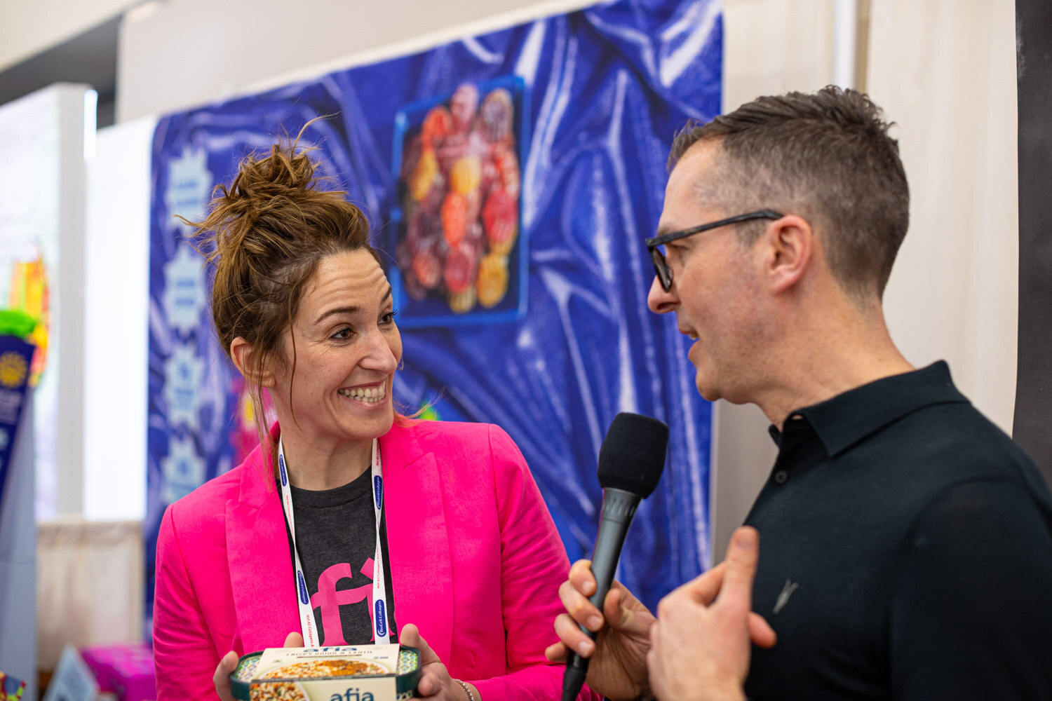 A smiling woman in a pink jacket holds a food package while an interviewer holds a microphone toward her