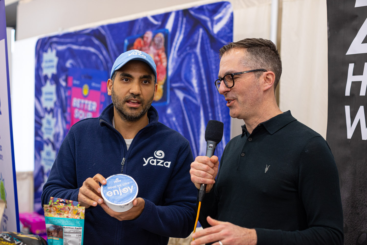 A man in a Yaza jacket holds a small tub while an interviewer with a microphone speaks with him at a booth with a purple backdrop