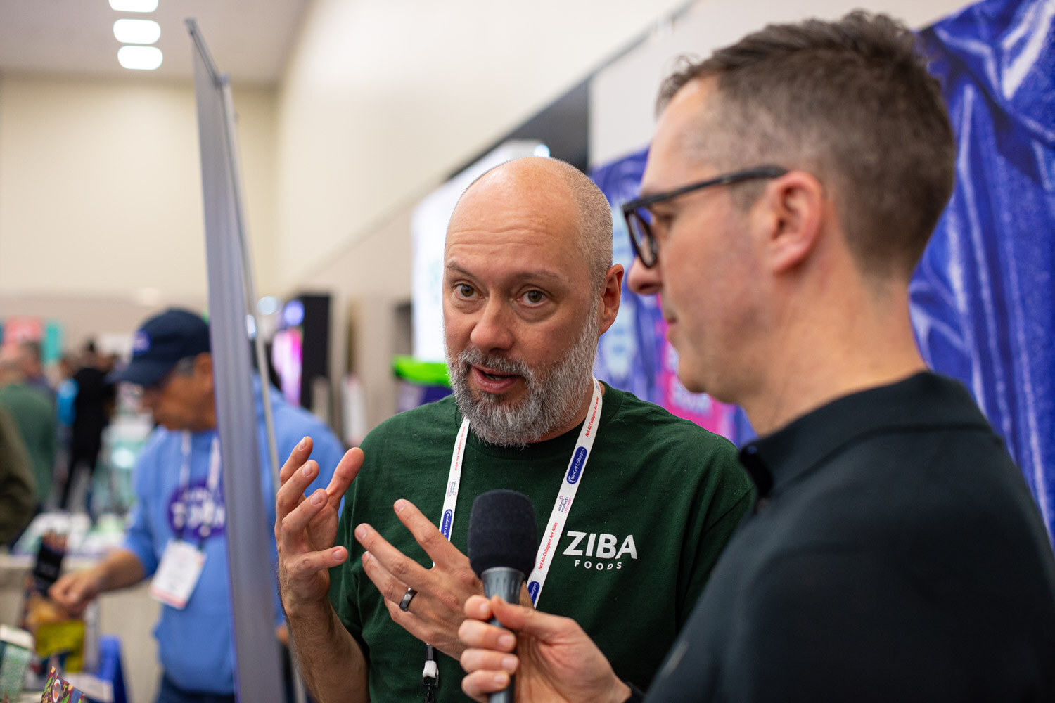 A man in a green Ziba Foods shirt speaks into a microphone held by an interviewer in a dark top at a booth