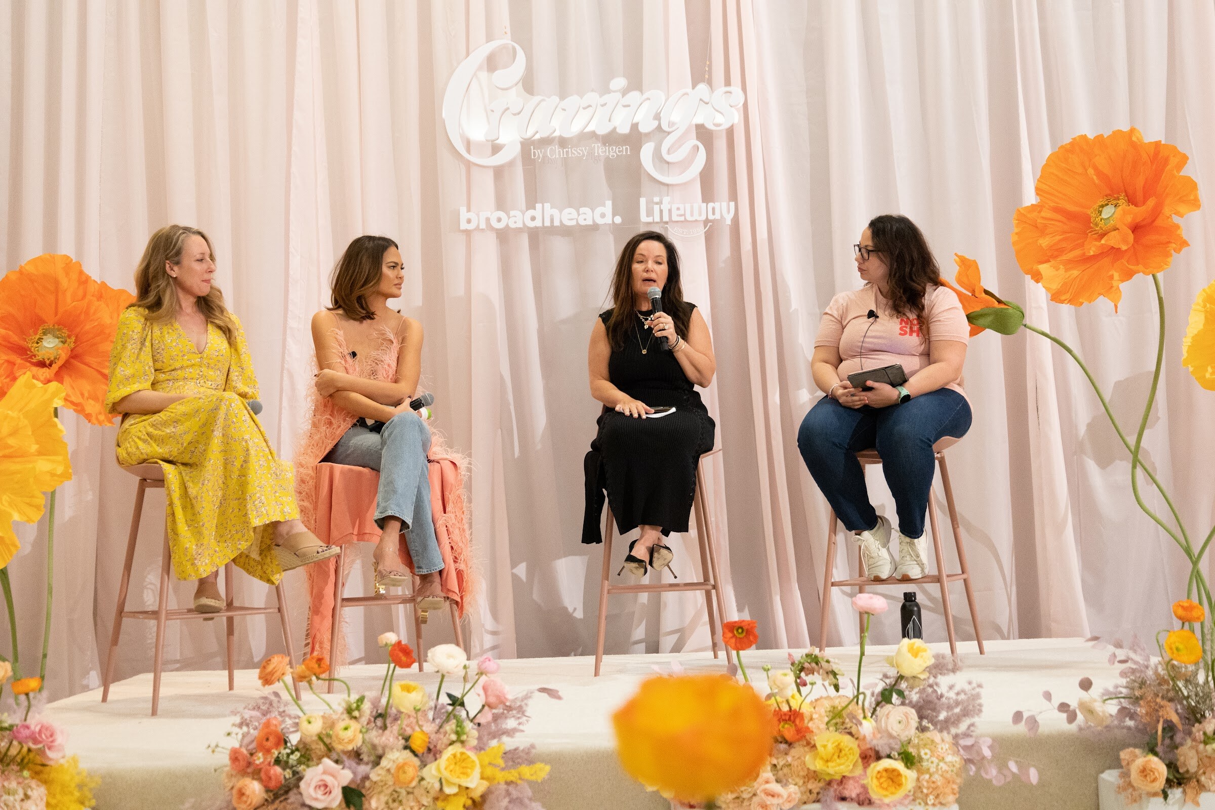 Four women seated on stools on a flower-decorated stage with pink curtains