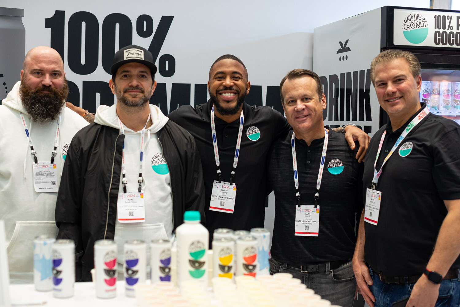 Five smiling men standing together behind a booth counter with bottles of drinks