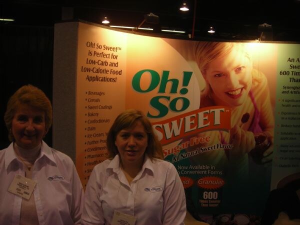 Two women in white shirts and name badges stand in front of an Oh! So Sweet sugar-free sweetener display showing text about low-carb applications and liquid and granular forms.