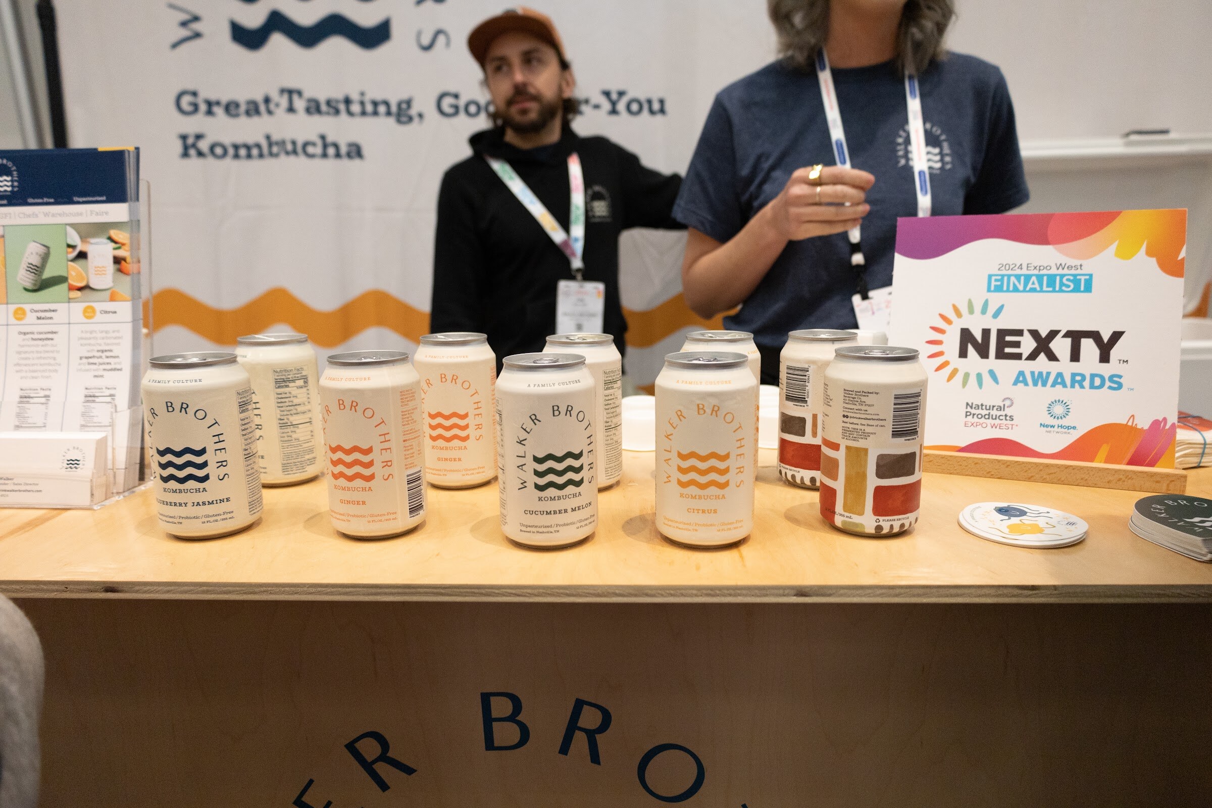 A booth counter lined with kombucha cans and a Nexty Awards Finalist sign, with two people behind it