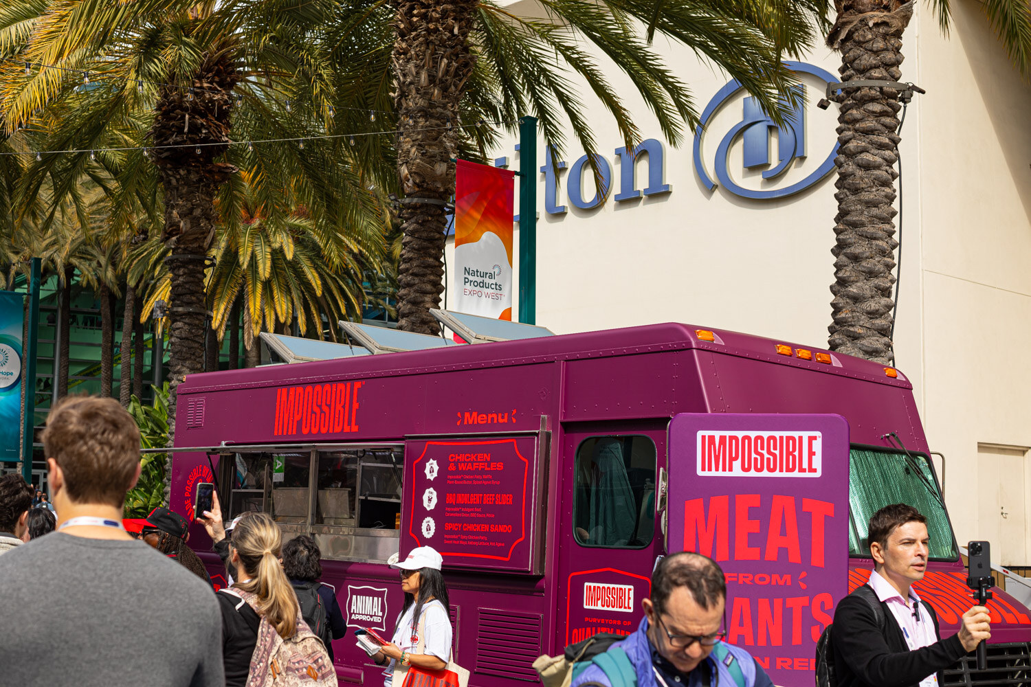 A purple Impossible food truck parked among palm trees with people gathered nearby