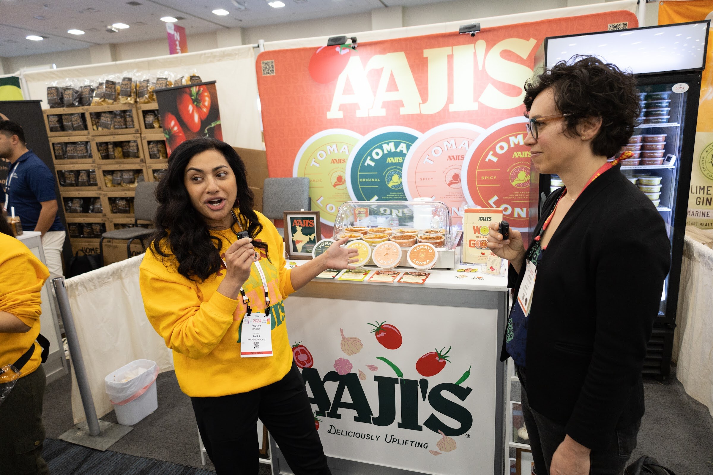 A woman in a yellow Aaji's sweatshirt speaks to another woman in front of the Aaji's tomato dip booth