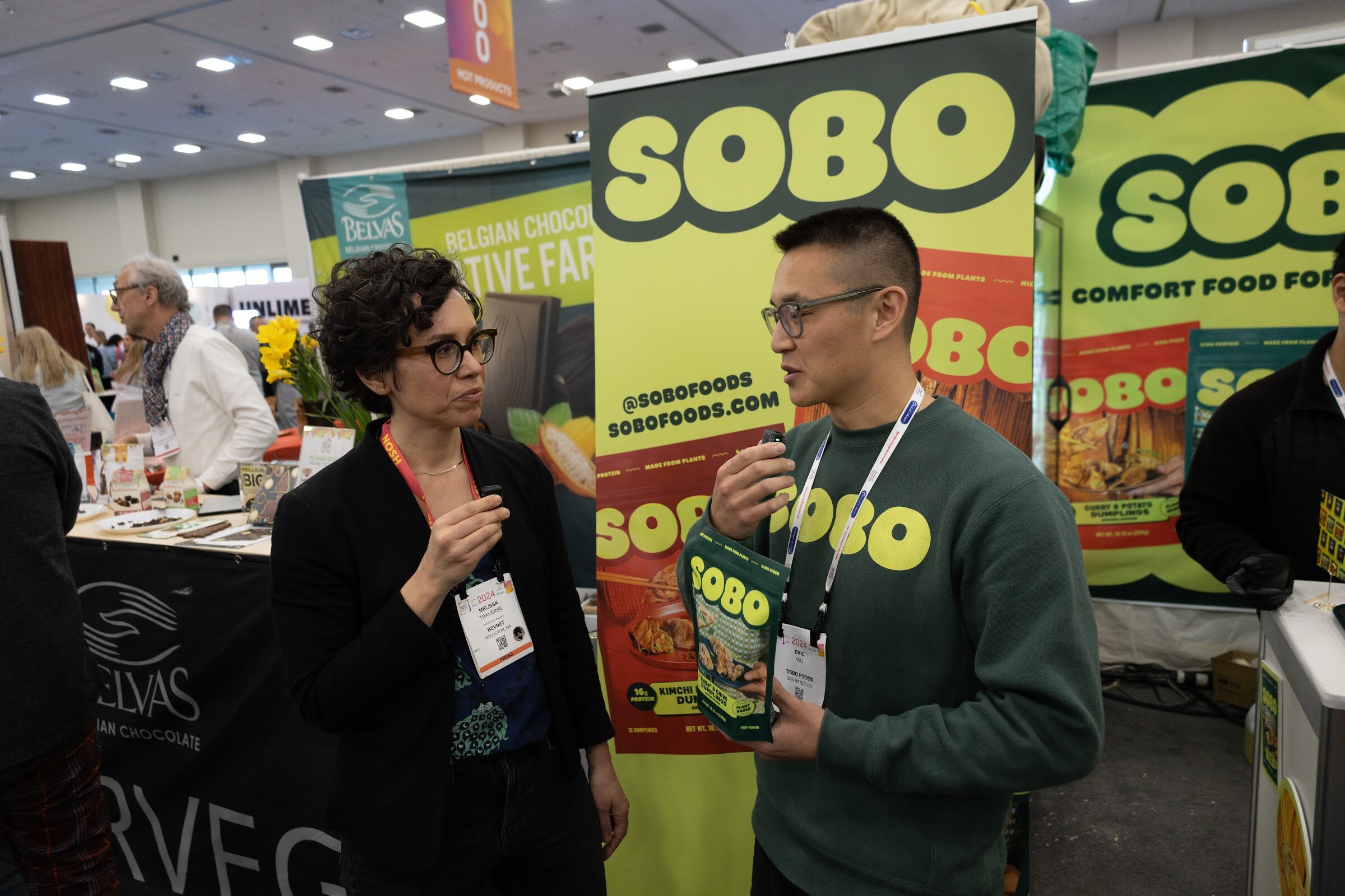 A woman and a man in a green sweatshirt talk in front of a large green SOBO sign on the show floor
