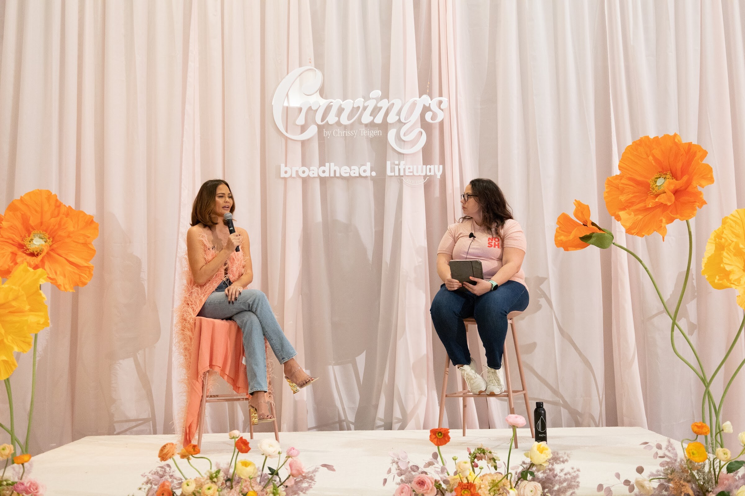 Two women seated on stools on a stage with pink curtains and orange flowers under a Cravings sign