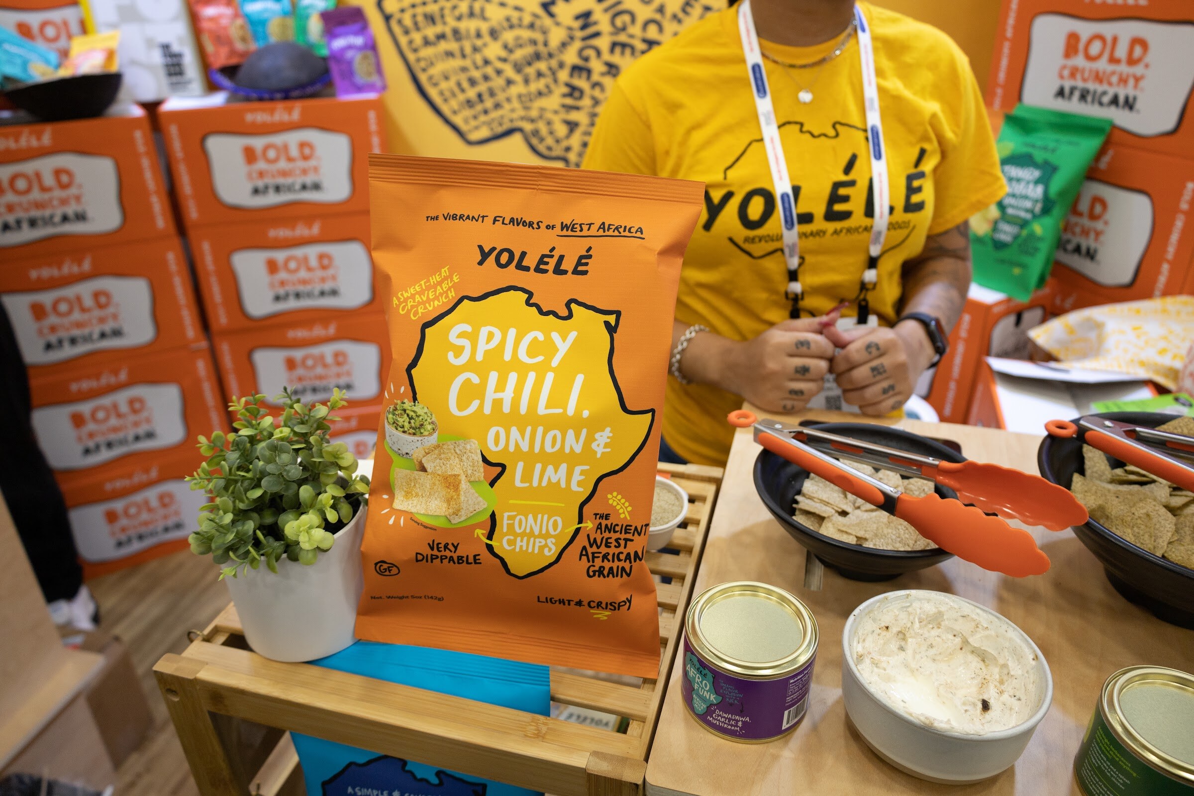Bags and tins of Yolele West African foods on a yellow table with a person in a yellow Yolele shirt behind