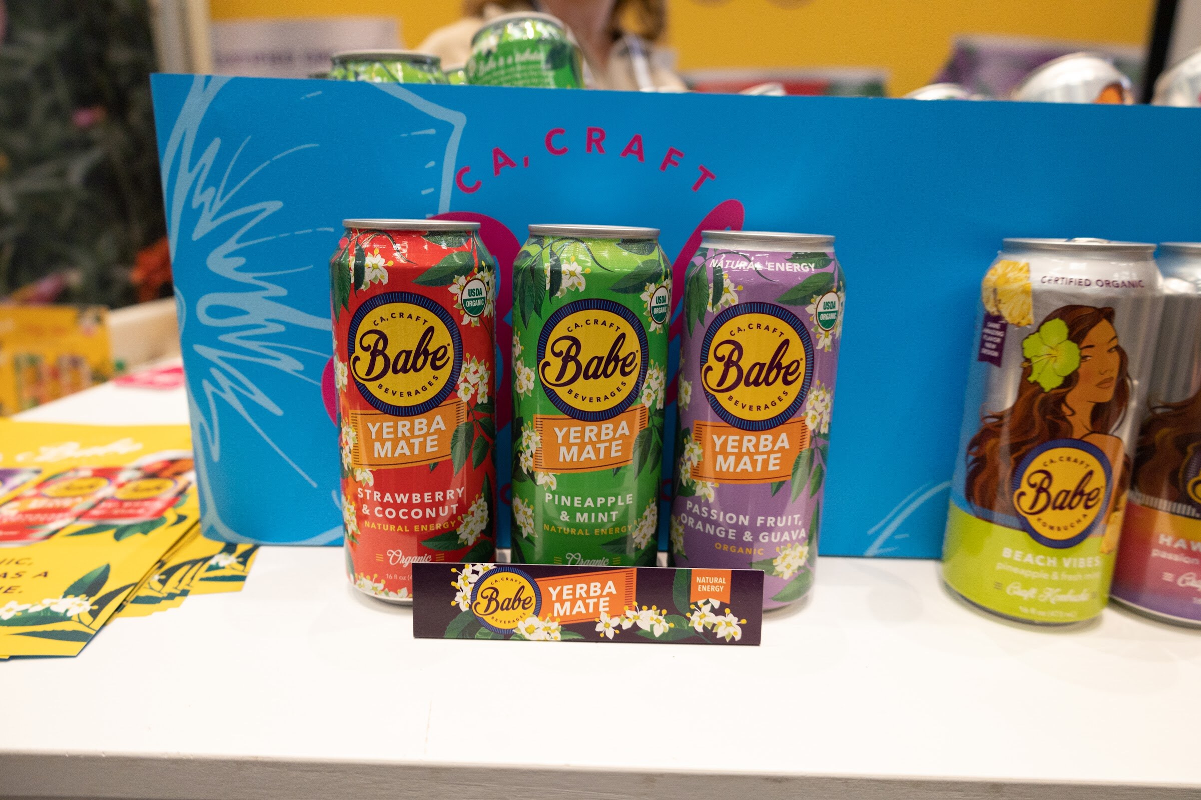 Babe yerba mate cans in several flavors on a blue display under a handwritten craft sign
