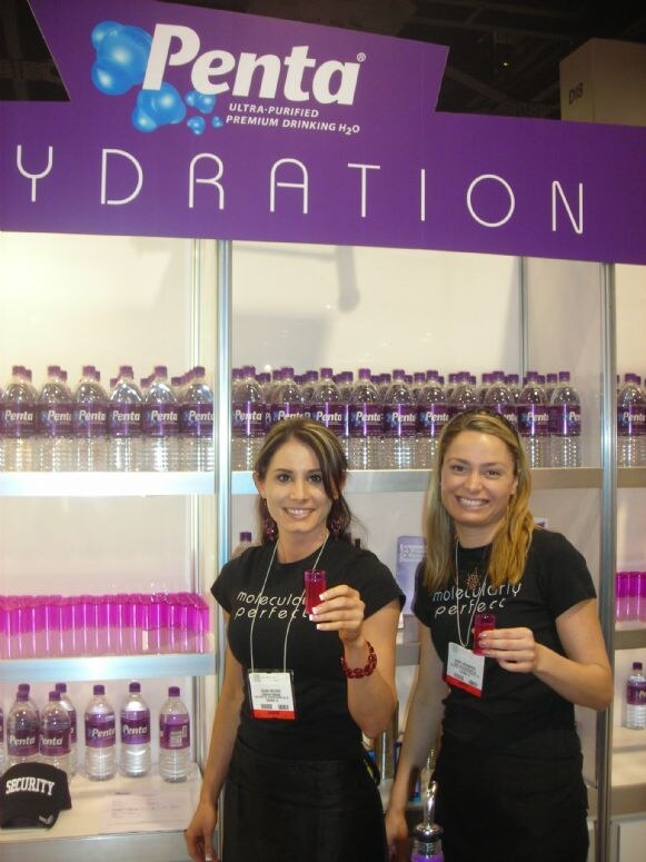 Two smiling women in black shirts hold small pink cups in front of shelves of Penta bottled water beneath a purple Penta Hydration sign.