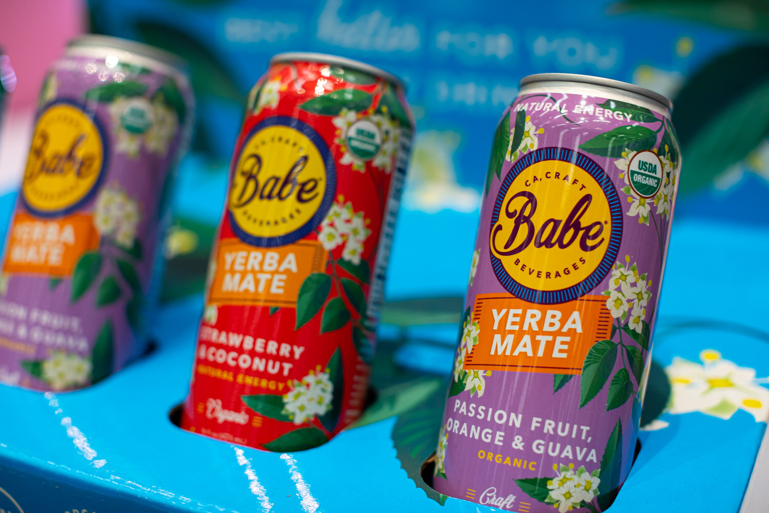 Three Babe yerba mate cans in strawberry coconut and other flavors on a blue display