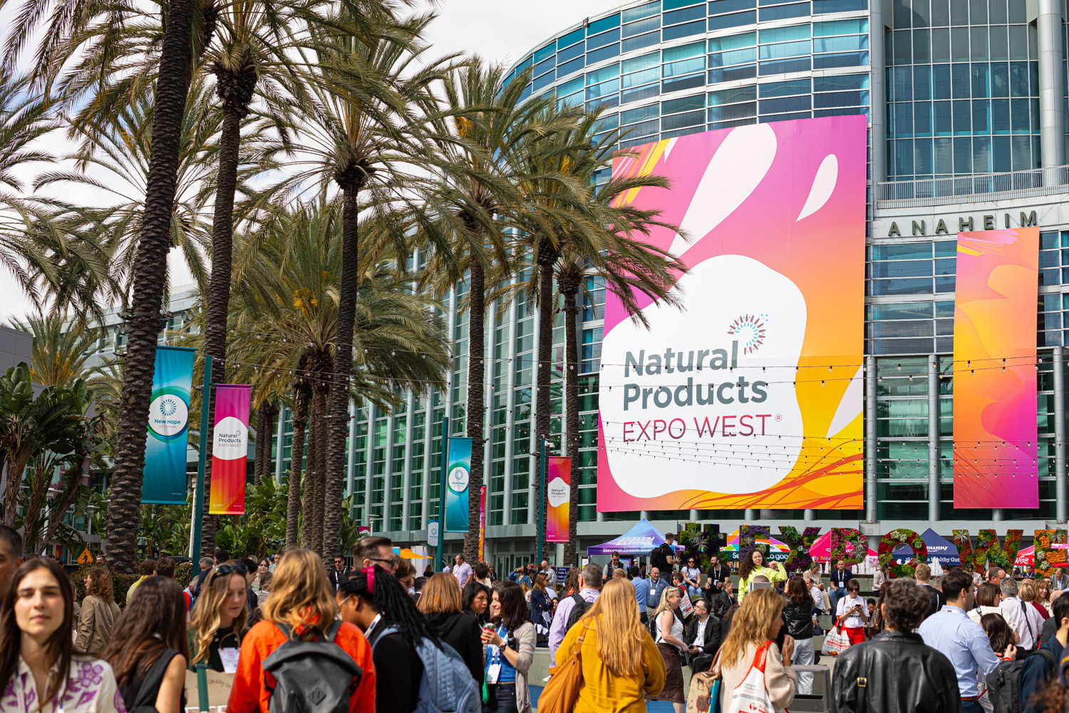 A crowd of attendees beneath palm trees and Natural Products Expo West banners on the convention center wall