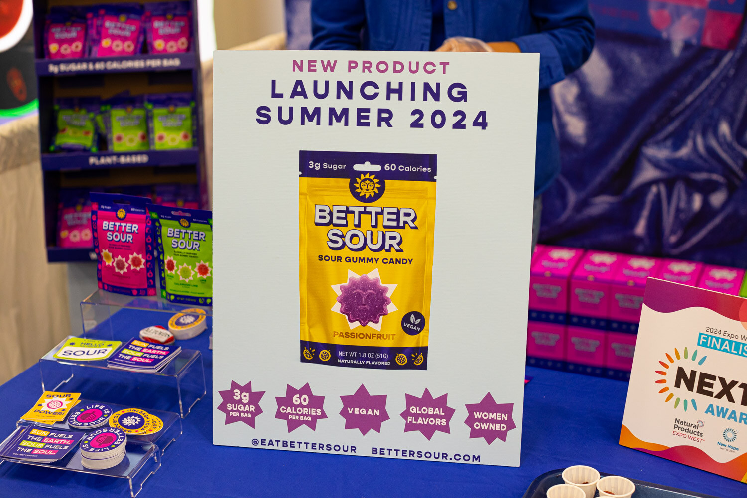 A Better Sour sour gummy candy pouch beside a New Product Launching Summer 2024 card and a Nexty Awards sign