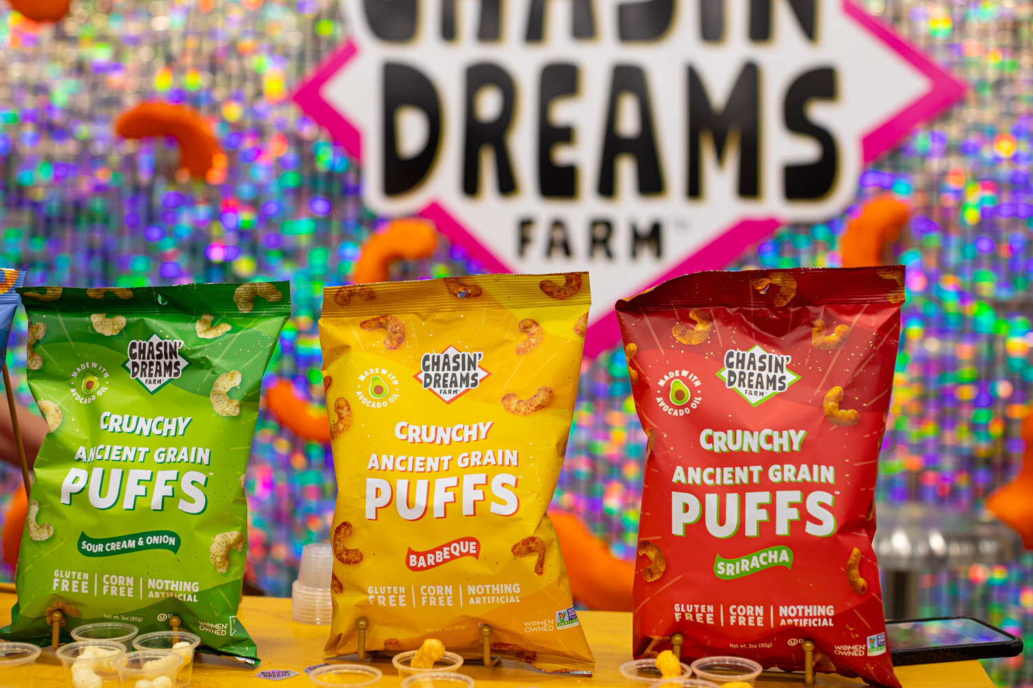 Three bags of Chasin' Dreams Farm crunchy ancient grain puffs in front of a glitter booth sign