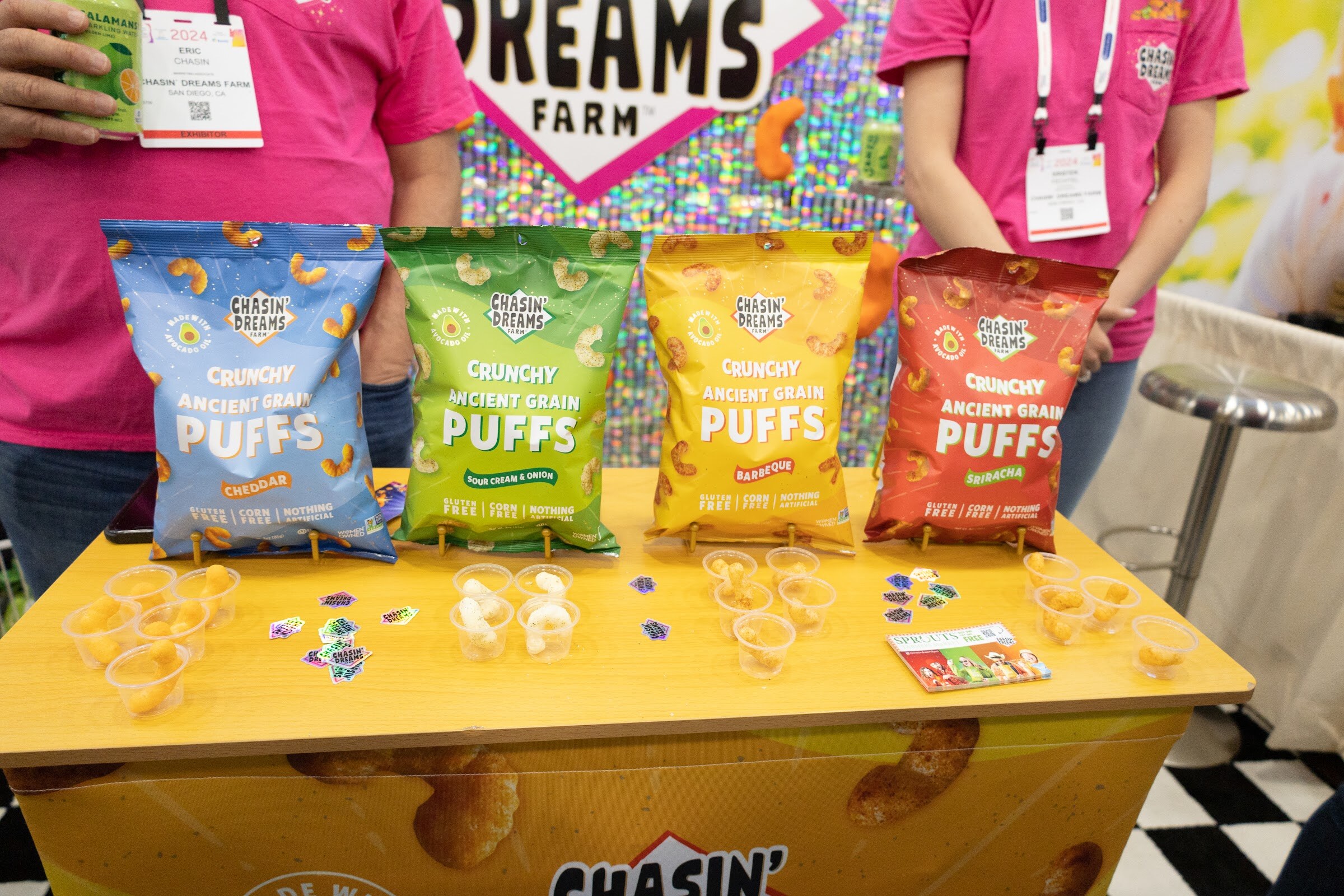 A yellow Chasin' Dreams Farm booth counter with four bags of ancient grain puffs and two people in pink shirts