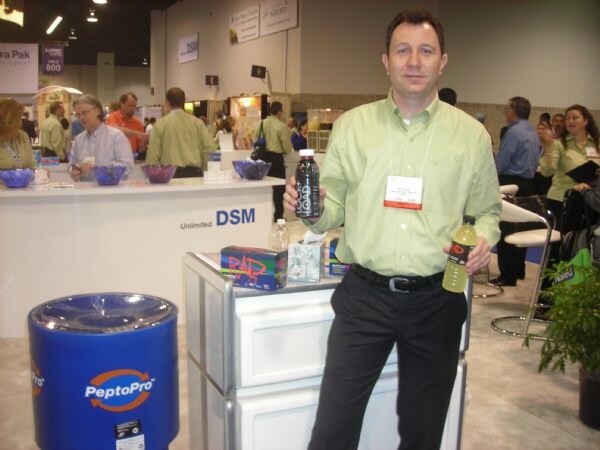 A man in a light green shirt holds a dark beverage bottle beside a blue PeptoPro drum and a DSM counter, with other attendees in the busy hall behind him.