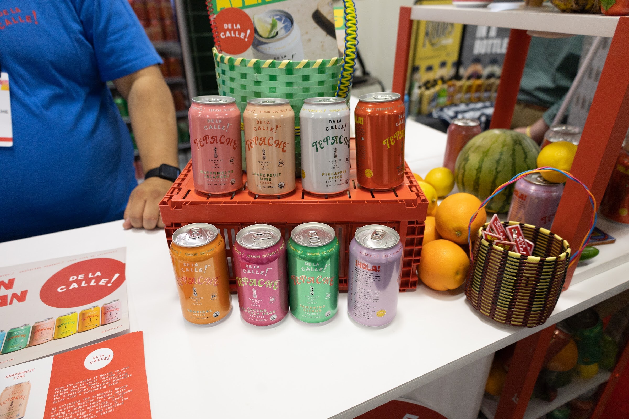 Several De La Calle cans on a red crate with fruit baskets and oranges behind them