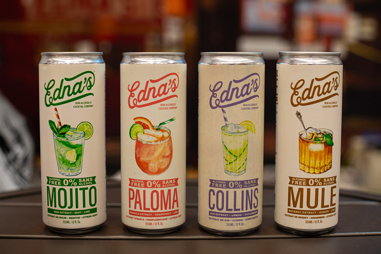 Four Edna's cans labeled Mojito, Paloma, Collins and Mule on a dark counter