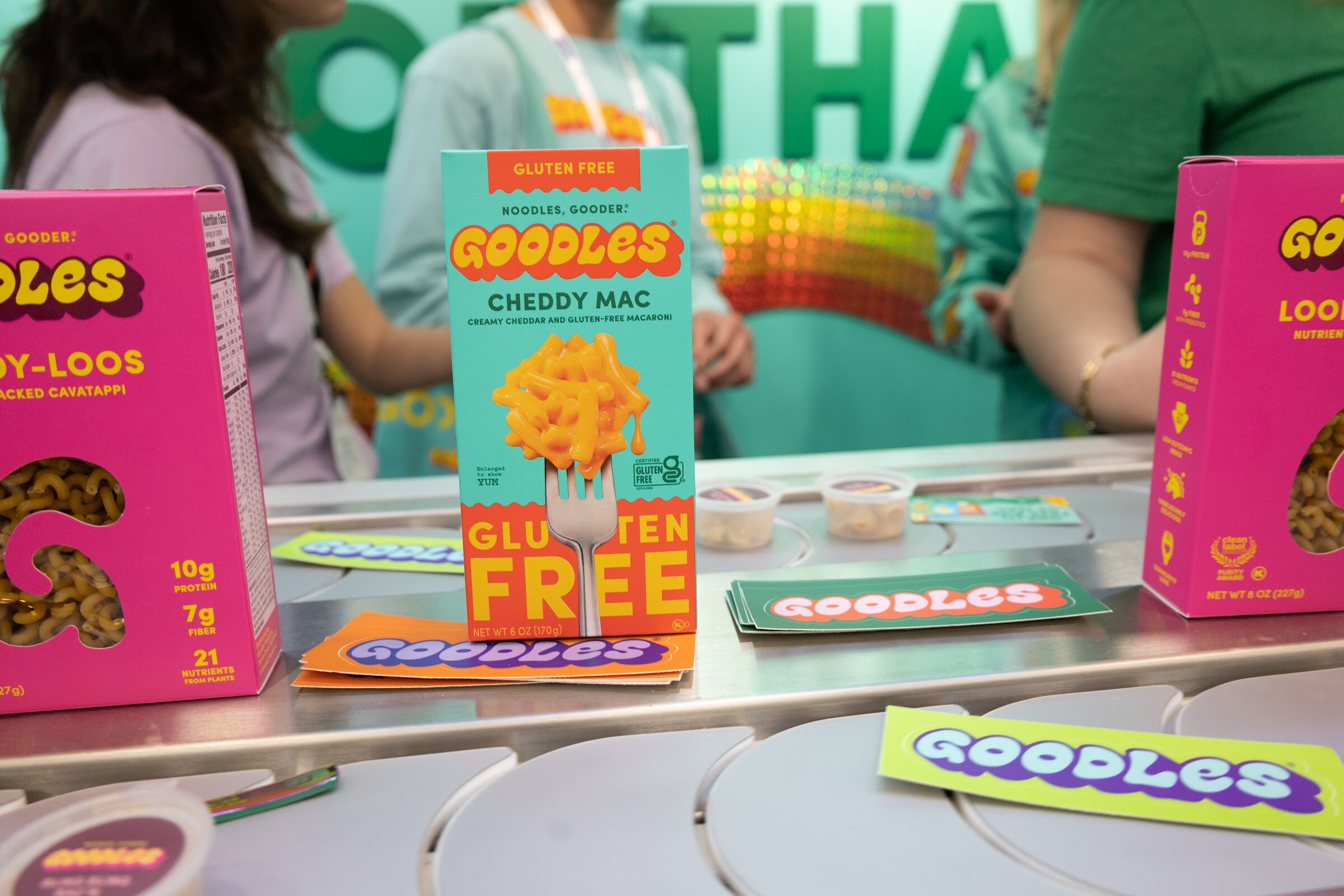 A Goodles cheddy mac box and gluten free packages on a metal counter at a teal Goodles booth