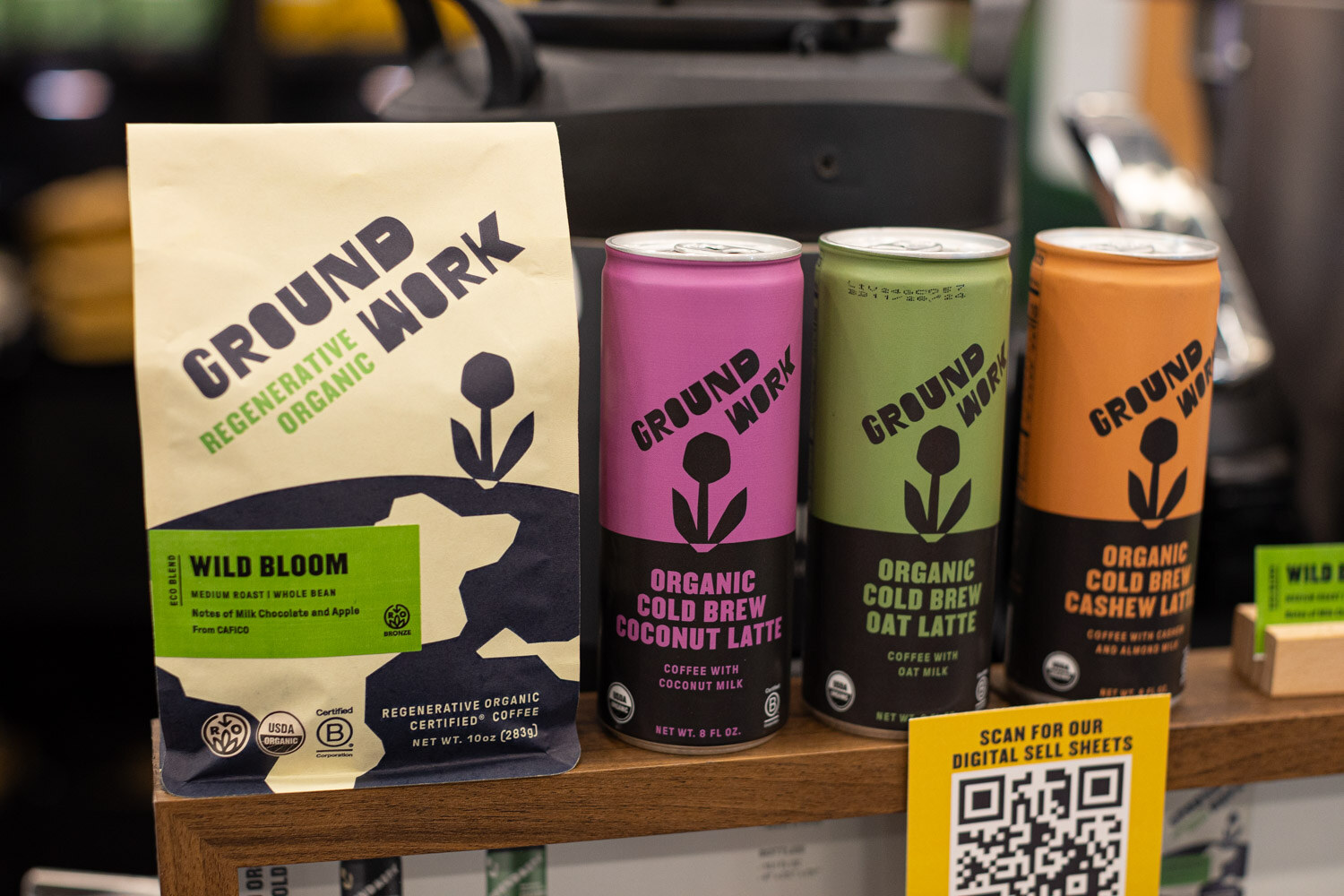 A bag of Ground Work Wild Bloom coffee next to three Ground Work cold brew cans on a wooden shelf