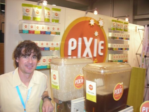 A smiling man with dark hair stands beside two beverage dispensers labeled Pixie Mate, with shelves of Pixie tea packages and a large orange Pixie sign behind him.