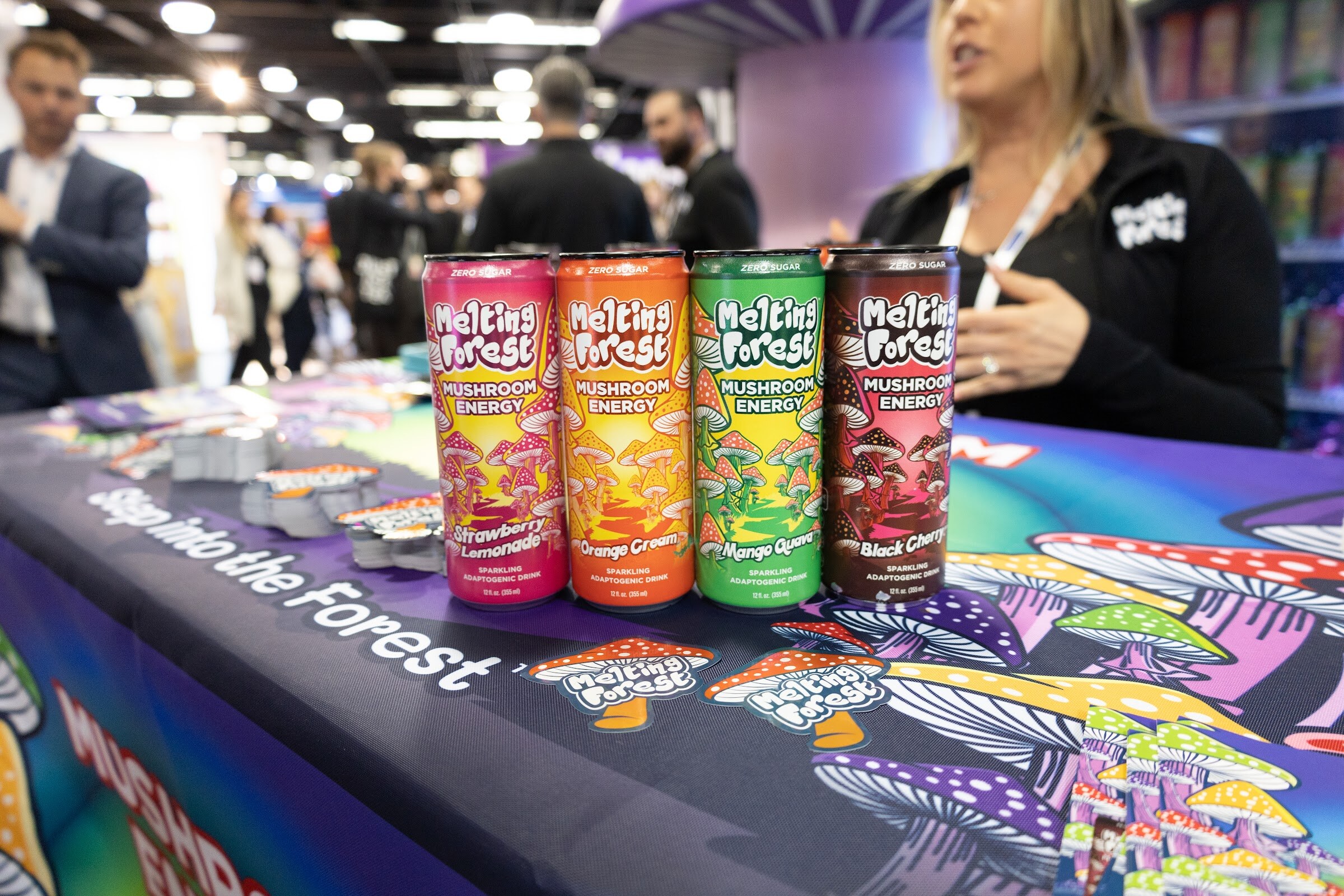 Four Melting Forest mushroom energy cans on a purple mushroom-print tablecloth at a booth