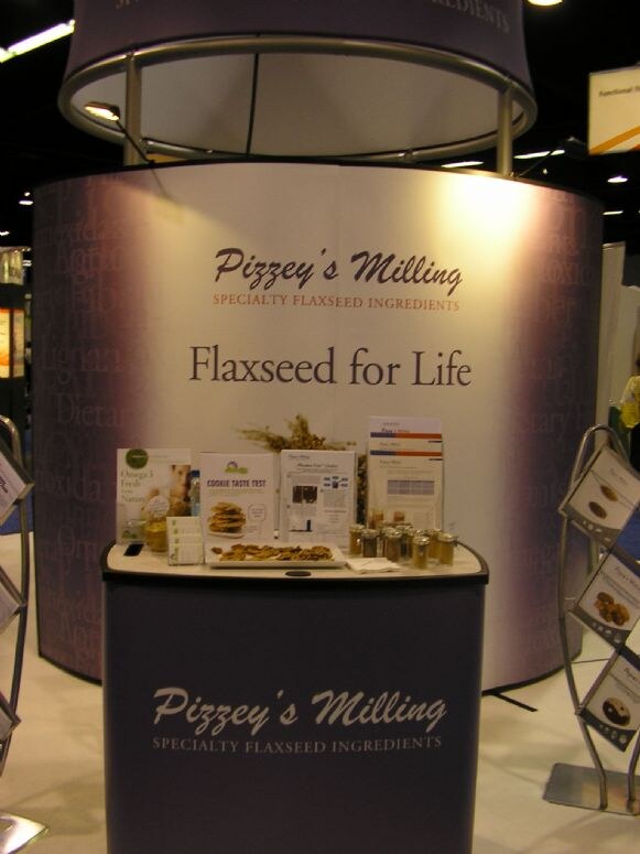 A curved Pizzey's Milling booth reading Flaxseed for Life, with a front counter holding brochures, a plate of cookies and small jars of flaxseed ingredients.