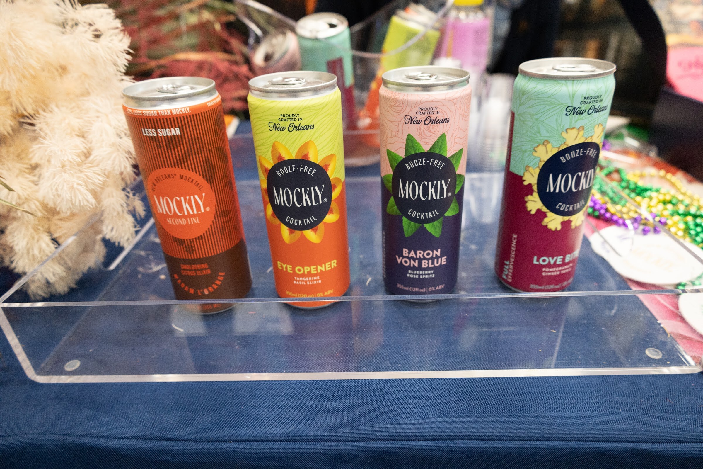 Four Mockly cans on a clear acrylic tray with Mardi Gras beads behind them