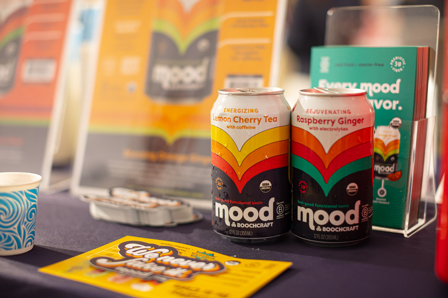 Two Mood sparkling drink cans, lemon cherry tea and raspberry ginger, on a dark table with brochures behind them