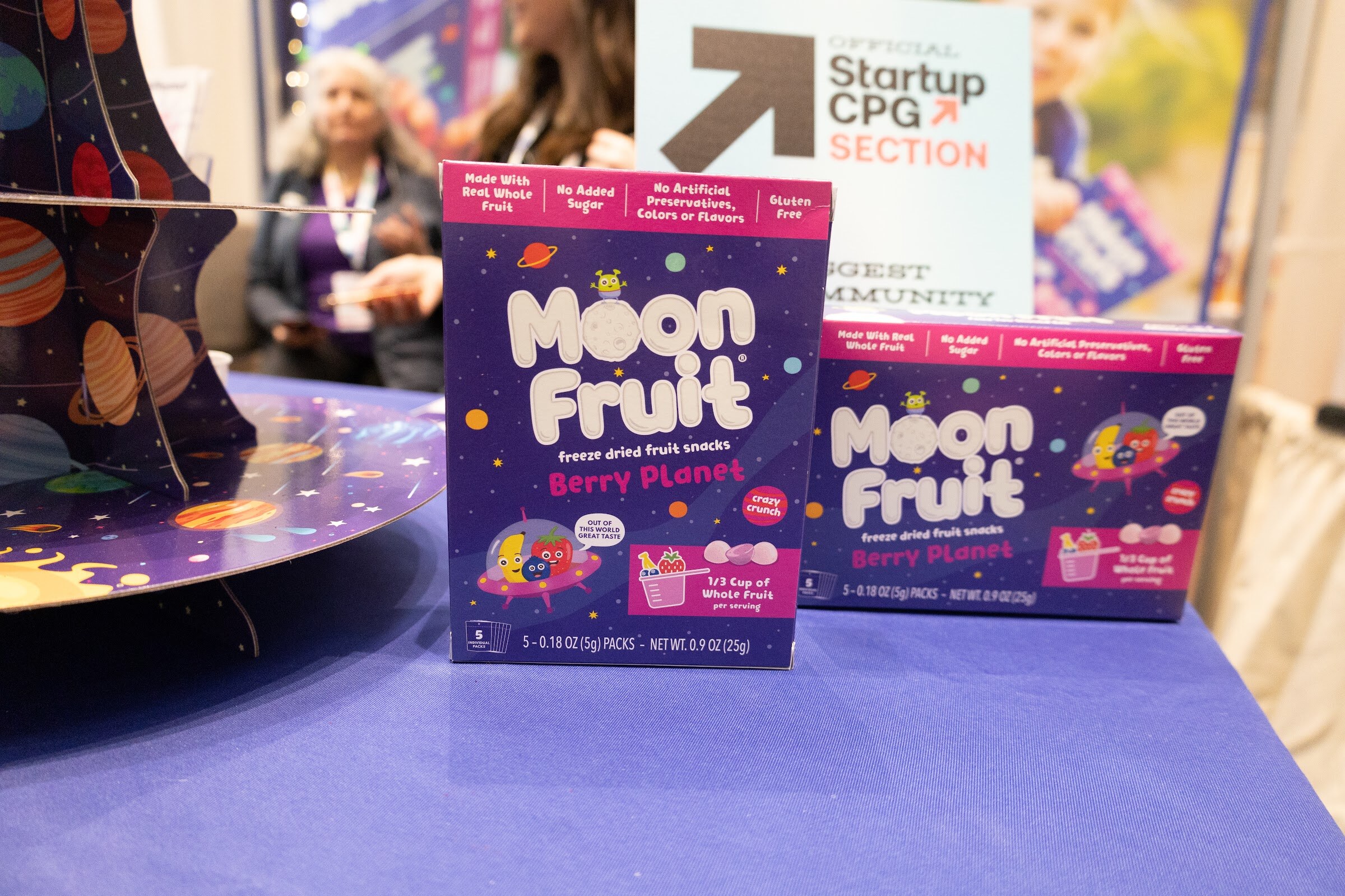 Moon Fruit freeze-dried fruit boxes on a purple tray in front of an Official Startup CPG Section sign