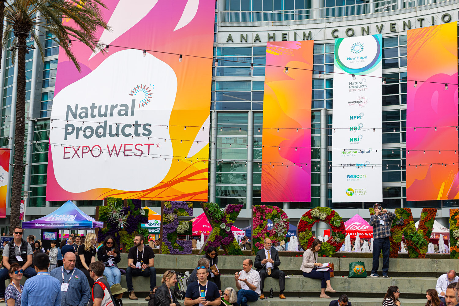 A large Natural Products Expo West banner on the Anaheim Convention Center above a floral display and seated attendees