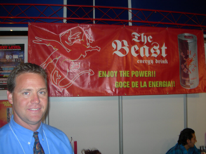 A smiling man in a blue shirt and patterned tie stands in front of a red The Beast energy drink banner with a winged creature, a can graphic and the text Enjoy the power.