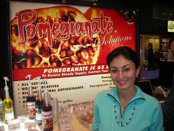 A smiling woman with glasses and a teal jacket stands in front of a Pomegranate Solutions sign showing pomegranate halves, with syrup bottles on the counter beside her.