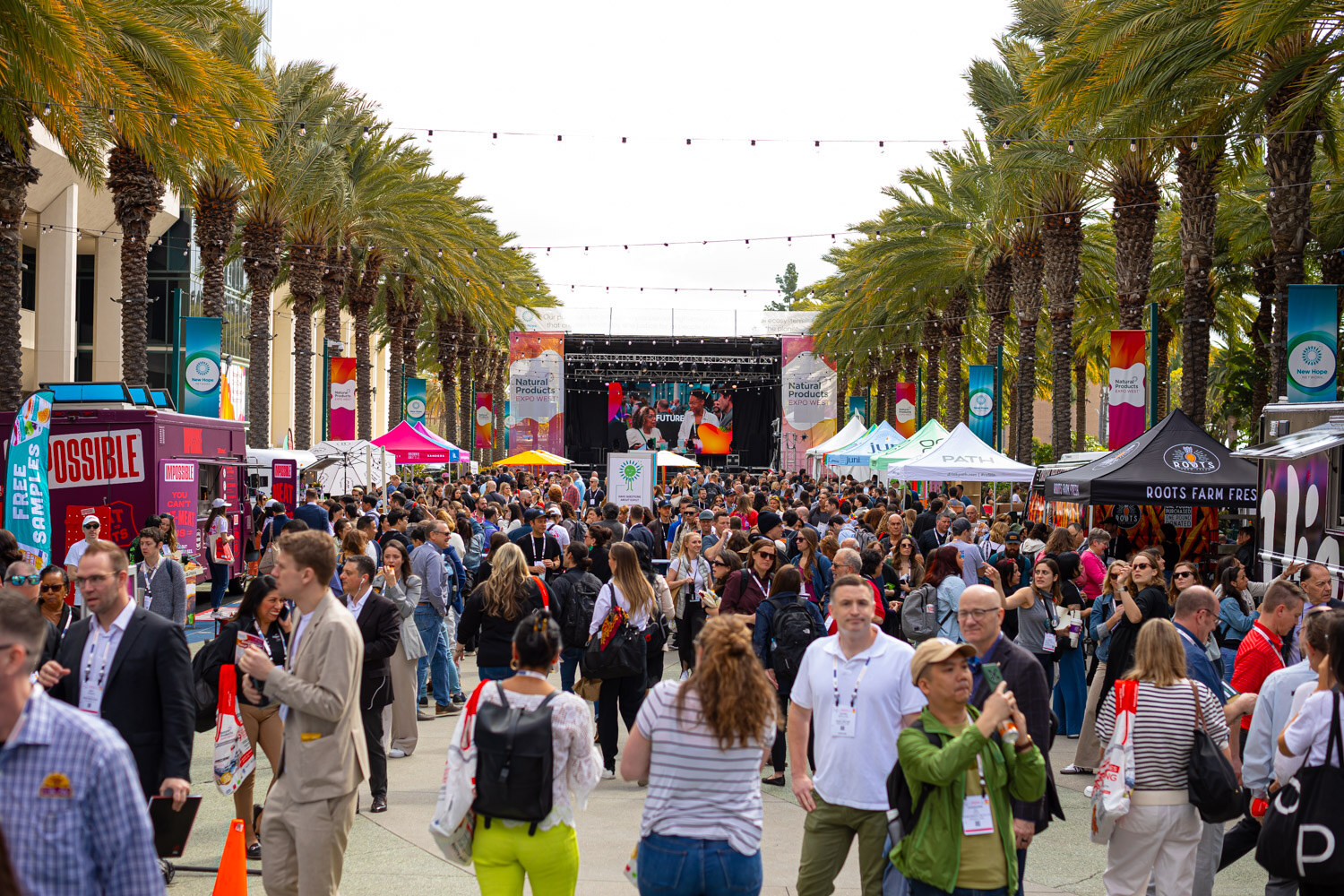 A busy outdoor plaza of attendees with food trucks and a stage beyond the palm trees