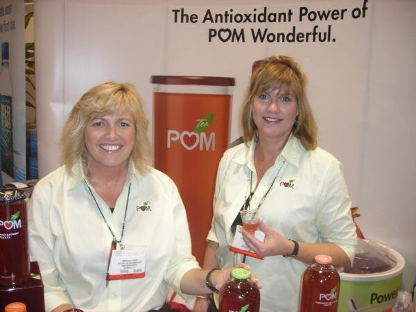 Two smiling women in cream shirts with POM logos stand at the POM Wonderful booth with bottles of POM Tea and juice in front of a sign about antioxidant power.