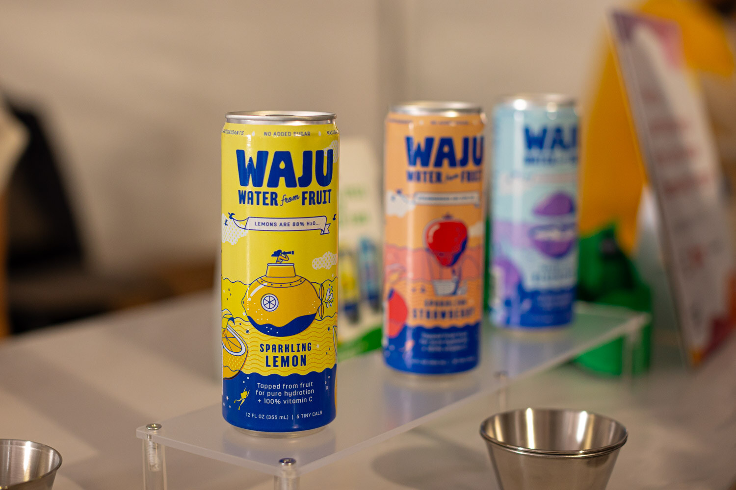 Three Waju water from fruit cans in lemon, strawberry and another flavor lined up on a clear stand