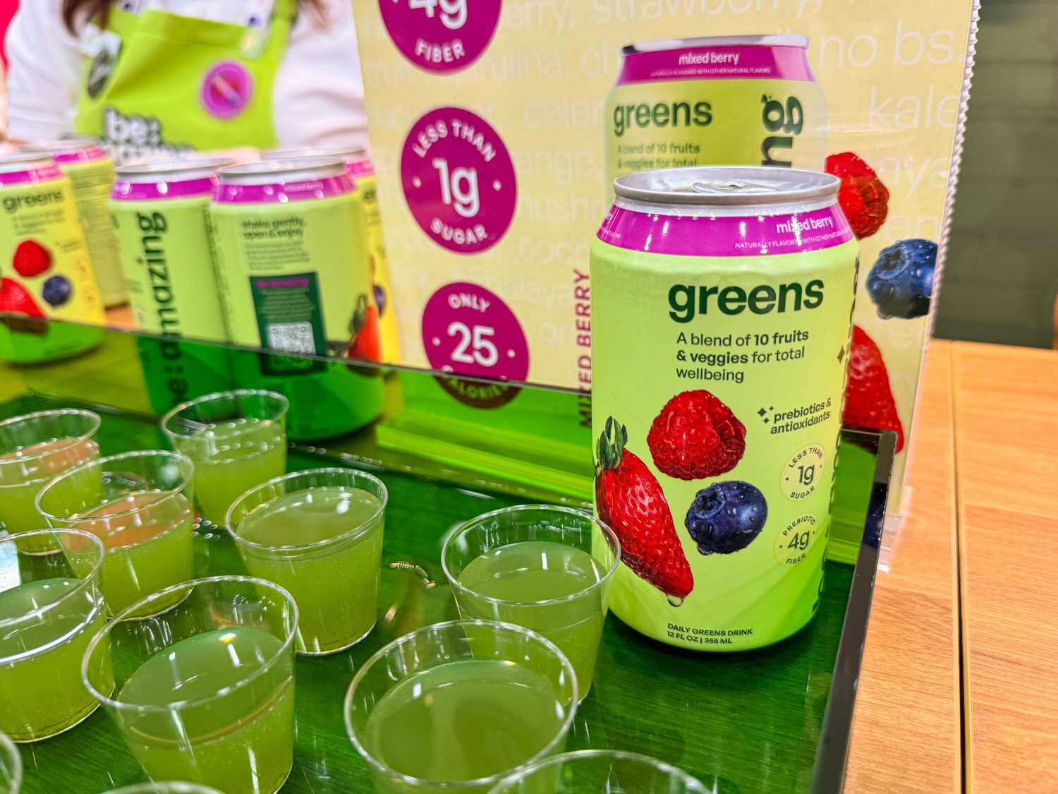 A green be:amazing greens can on a green display with sample cups of green drink