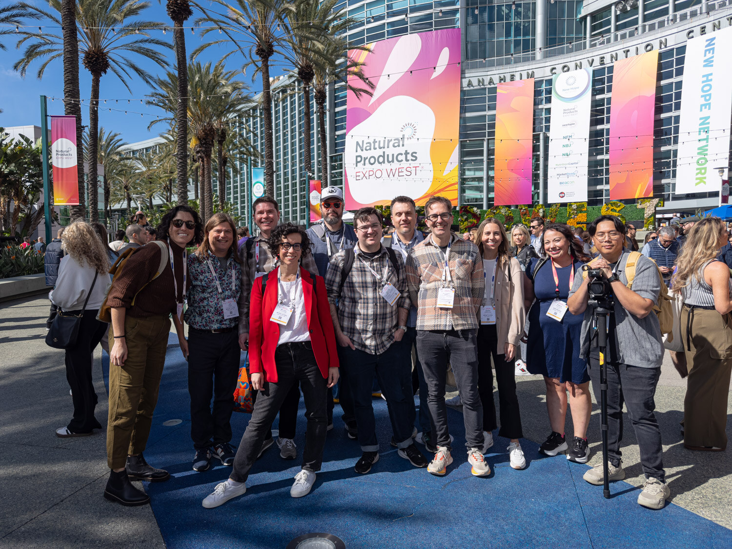 A group of about a dozen people posing in front of Natural Products Expo West banners with a person holding a camera on a tripod