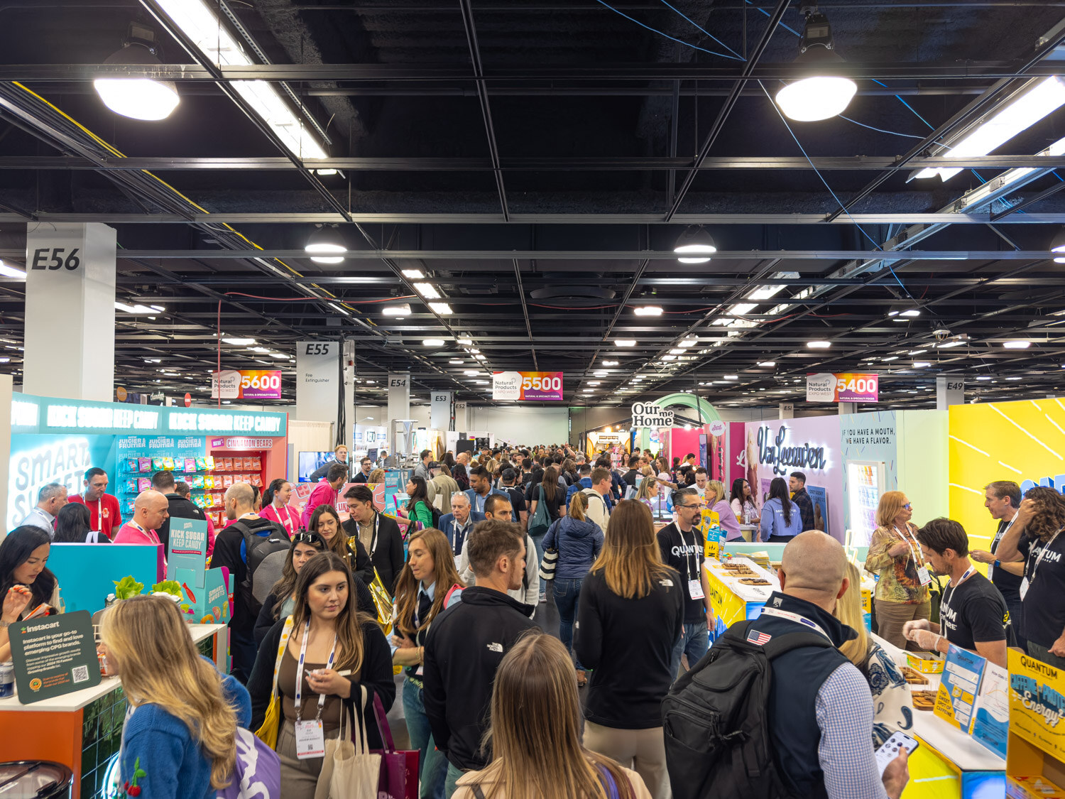 A crowded aisle in the exhibit hall under a high ceiling with colorful booths on both sides